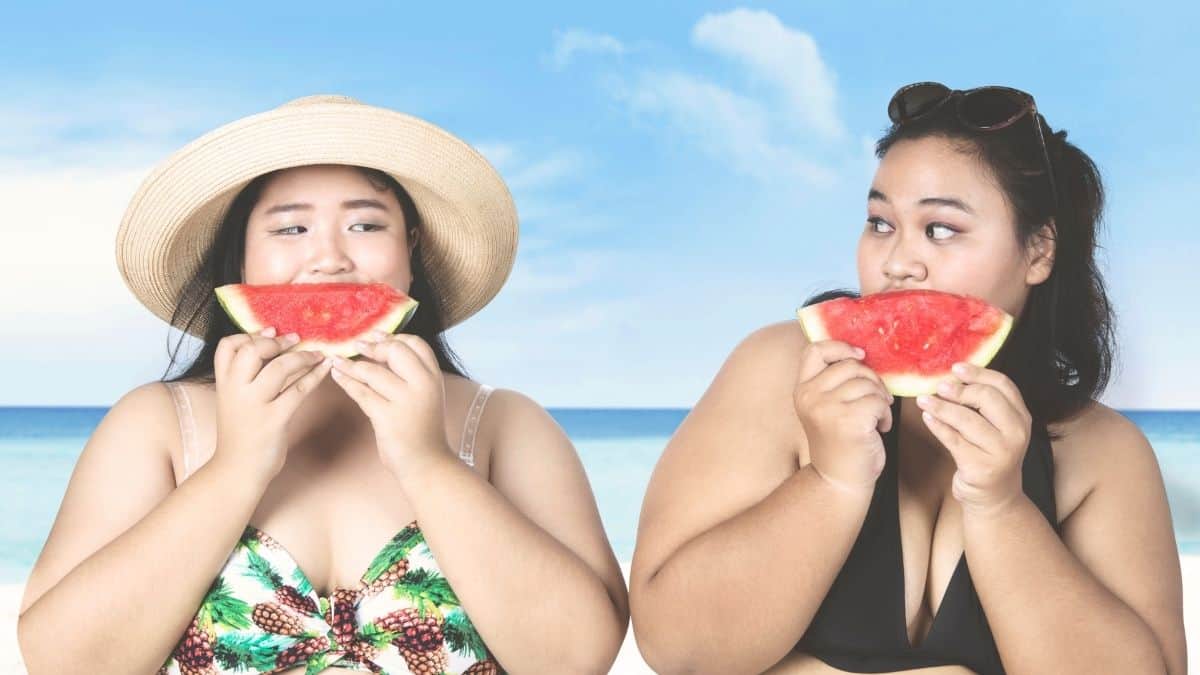 two overweight women in swimsuits sitting on the beach eating watermelon.
