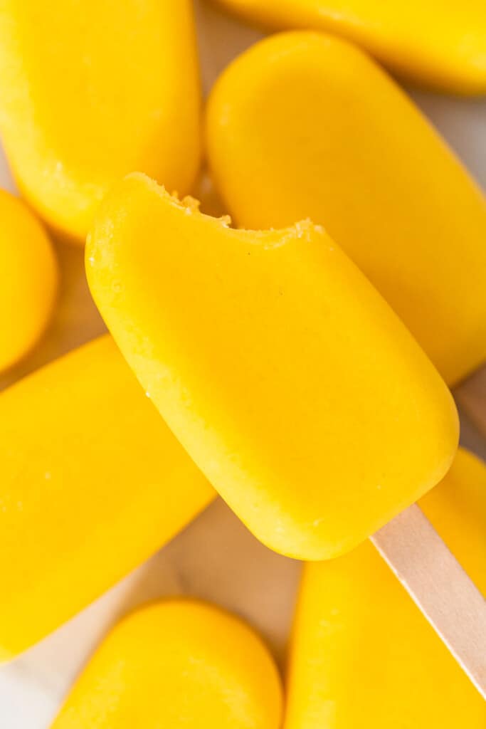Mango pops on a white counter with one on top with a bite taken out of it,