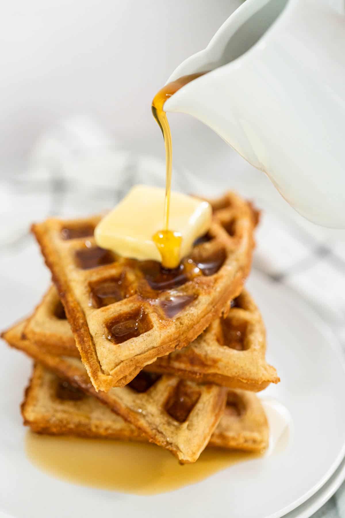 A stack of Oat Flour Waffles with a pat of butter on top and syrup being poured from a white container.