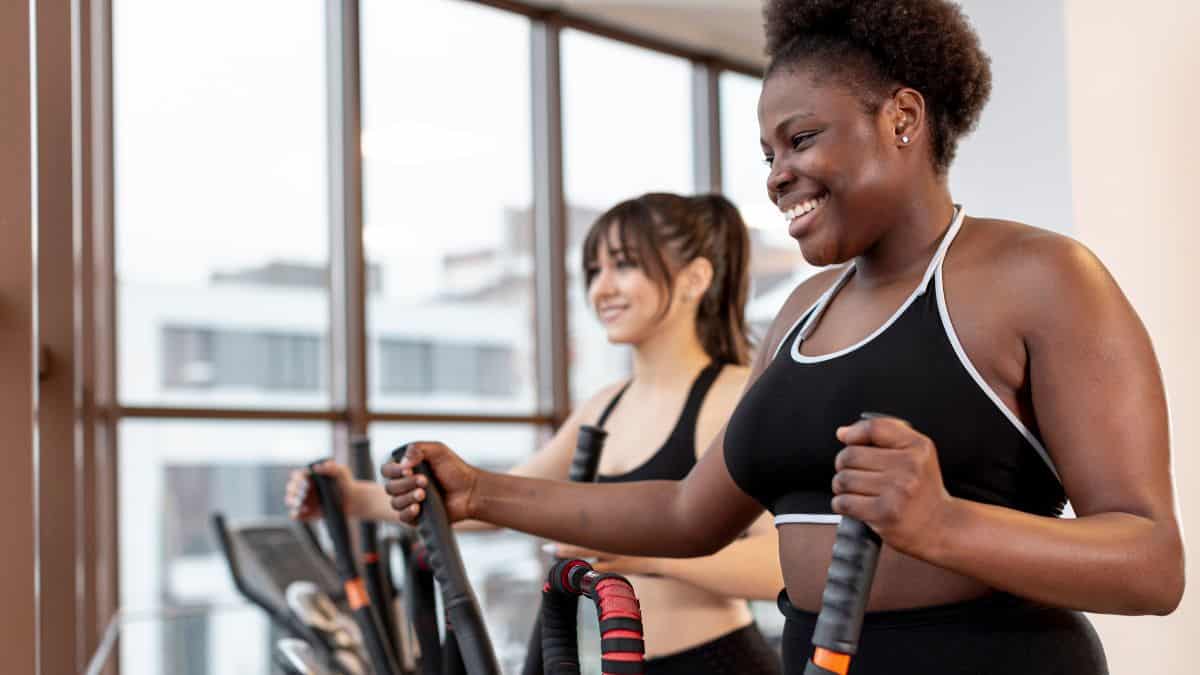 a smiling woman with short curly hair using an elliptical machine with another woman in the background.