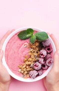 a bowl of yogurt decorated with granola, frozen raspberries and mint on a pink background.