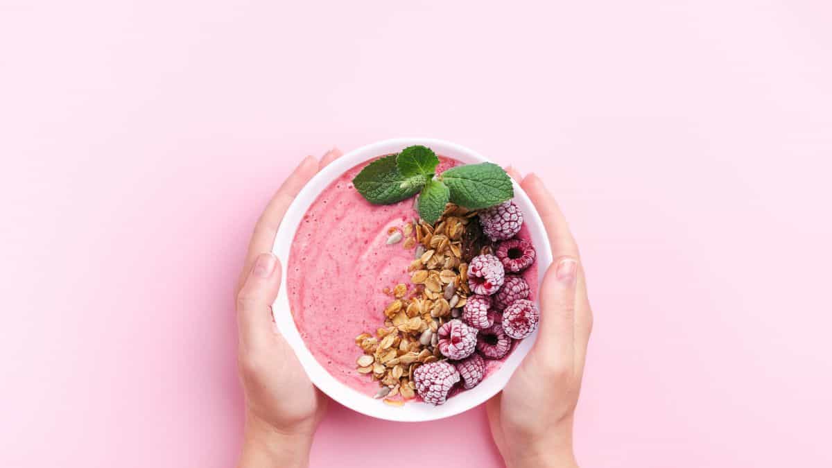 a bowl of yogurt decorated with granola, frozen raspberries and mint on a pink background.