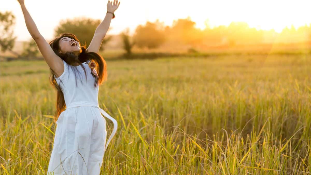 woman smiling and jumping with arms wide in field on sunny day.