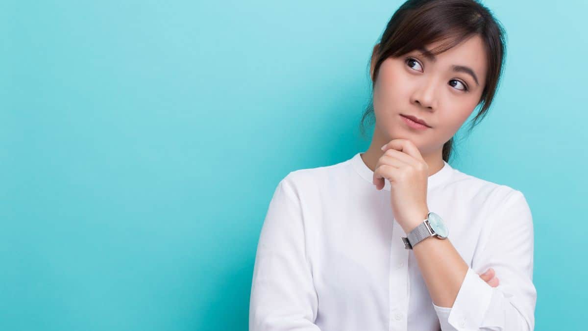 a woman with brown hair pulled back stands against a blue wall thinking.