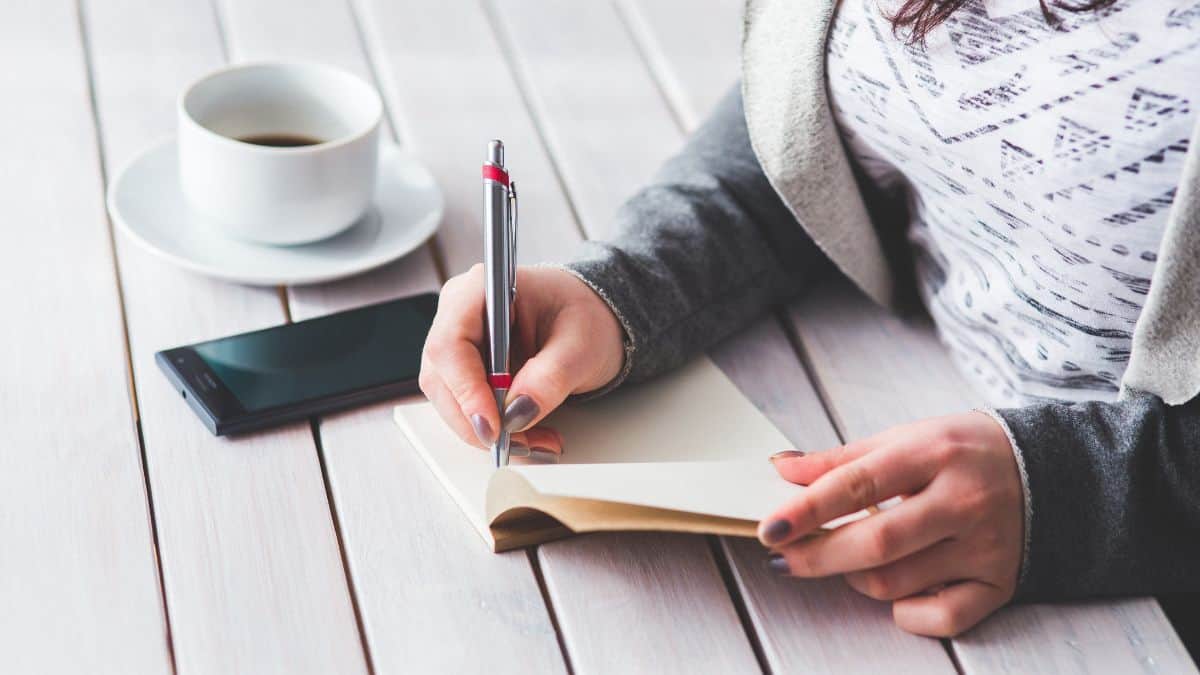 woman writing in a journal at the table with cell phone and coffee cup next to her.